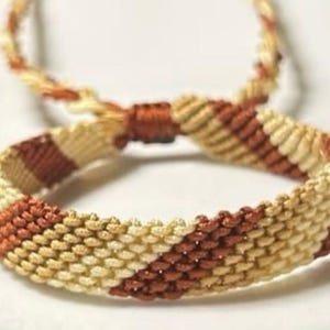 May include: A handmade macrame bracelet in shades of tan and brown. The bracelet features a woven pattern with diagonal stripes. The adjustable closure is also macrame. This accessory is perfect for adding a touch of bohemian style.