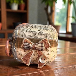 May include: A clear, decorative case for small electronics, featuring a brown and cream floral pattern. The case is adorned with a brown bow and a button closure. The case has a handle on the side.