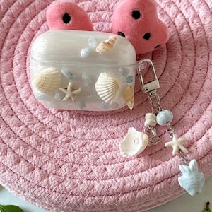 May include: A clear AirPods case decorated with seashells, starfish, and small blue stones. The case has a silver clasp and a charm with a seashell, starfish, and a white bunny. Two pink fuzzy heart-shaped objects are behind the case.