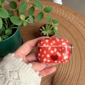 May include: A red and white polka-dot AirPod case with a heart-shaped graphic. The case is held in a hand, with a green plant and a small succulent in the background. The case has a glossy finish.