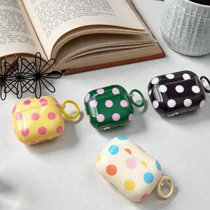 May include: Four colorful AirPod cases with polka dot patterns. The cases are yellow with pink dots, green with pink dots, black with white dots, and off-white with multi-colored dots. Each case has a matching colored ring for attachment.