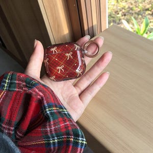 May include: A red plaid AirPod case with a gold bow pattern. The case has a clear, glossy finish and a gold-colored ring for attachment. The case is held in a hand, with a red and green plaid shirt sleeve visible in the foreground.