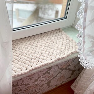May include: A beige, hand-knitted rectangular mat with a textured, woven pattern. The mat is placed on a windowsill, next to a window and sheer curtains. The mat's neutral color complements the window's white frame.
