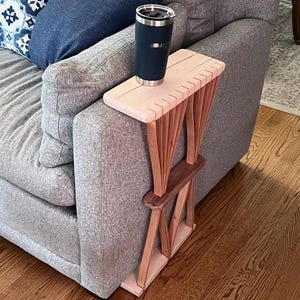 Puede incluir: Una mesa de brazo de sofá de madera con una parte superior de color claro y una base de color oscuro. La mesa está diseñada para encajar sobre el brazo de un sofá. Un vaso térmico azul oscuro está sobre la mesa.