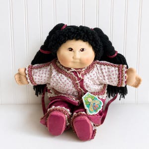 May include: A Cabbage Patch Kids doll with black yarn hair styled in pigtails with maroon ribbons. The doll wears a white quilted jacket with a small floral pattern and maroon trim, paired with matching maroon satin pants and a paper hang tag.