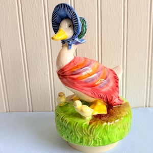 May include: A ceramic figurine of a white duck wearing a blue bonnet and a multi-colored shawl, standing on a green grassy base with two small yellow ducklings. The duck has a bright yellow beak and the shawl features stripes of red, orange, and purple.