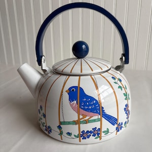 May include: White enamel teapot with a blue bird design. The teapot features a blue handle and knob, and a bird illustration inside a cage design. Floral accents in blue and green decorate the sides. The teapot is a charming kitchenware item.