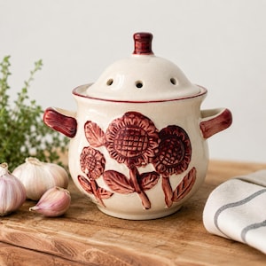 May include: Cream-colored ceramic garlic keeper with a lid, featuring a sunflower design in a deep red hue. The keeper has two handles and a small knob on the lid. Garlic bulbs are visible on a wooden surface, suggesting kitchen storage.