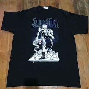 May include: Black t-shirt with the name "Austin" in blue, and a graphic of a skeleton holding a snake. The skeleton is white and grey, with a blue and white background. The shirt is made of a soft material.