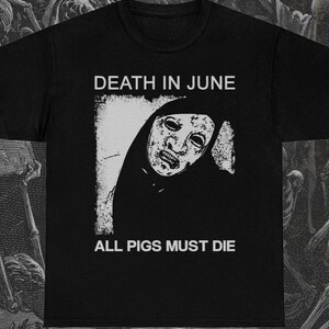 Può includere: T-shirt nero con grafica bianca di una figura mascherata e la scritta "DEATH IN JUNE" sopra e "ALL PIGS MUST DIE" sotto. Il design è un'immagine ad alto contrasto.