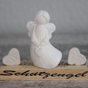 Puede incluir: Una figura de ángel blanco sosteniendo un corazón, flanqueada por dos decoraciones en forma de corazón. El ángel y los corazones están sobre una base de madera con la palabra "Schutzengel" en escritura negra. El fondo es gris neutro.