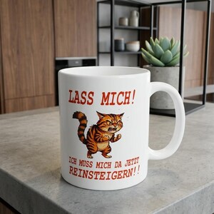 Könnte beinhalten: Weißer Keramikbecher mit Henkel, mit einer Cartoon-Illustration einer wütenden orangefarbenen Tabby-Katze. Der Becher trägt den deutschen Text "Lass mich!" und "Ich muss mich da jetzt reinsteigern!!" in Rot.