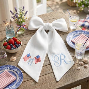 Könnte beinhalten: Ein weißes Tischband in Schleifenform mit gestickten amerikanischen Flaggen und einem blauen Initialen. Das Band liegt auf einem Holztisch mit patriotischen Dekorationen, darunter eine Schüssel mit Beeren und Gläsern Limonade.