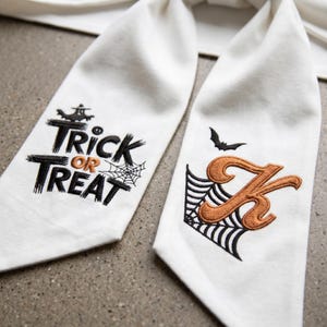 May include: Two white Halloween-themed fabric napkins. One napkin has the words "Trick or Treat" embroidered in black and orange, with a spiderweb design. The other napkin has an embroidered orange letter "K" with a spiderweb and a bat.