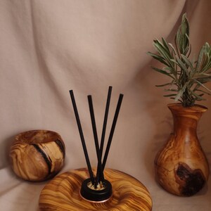 Puede incluir: Un difusor de aceites esenciales de madera con varillas negras, un pequeño jarrón de madera con vegetación y un pequeño cuenco de madera. El difusor tiene un borde color cobre. Los artículos son de madera con veta natural.