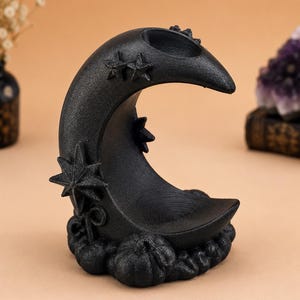 May include: A black crescent moon-shaped incense holder with a textured, glittery finish. The piece features raised star motifs on the curve and a base decorated with small, sculpted pumpkin shapes. A circular indentation sits at the top.