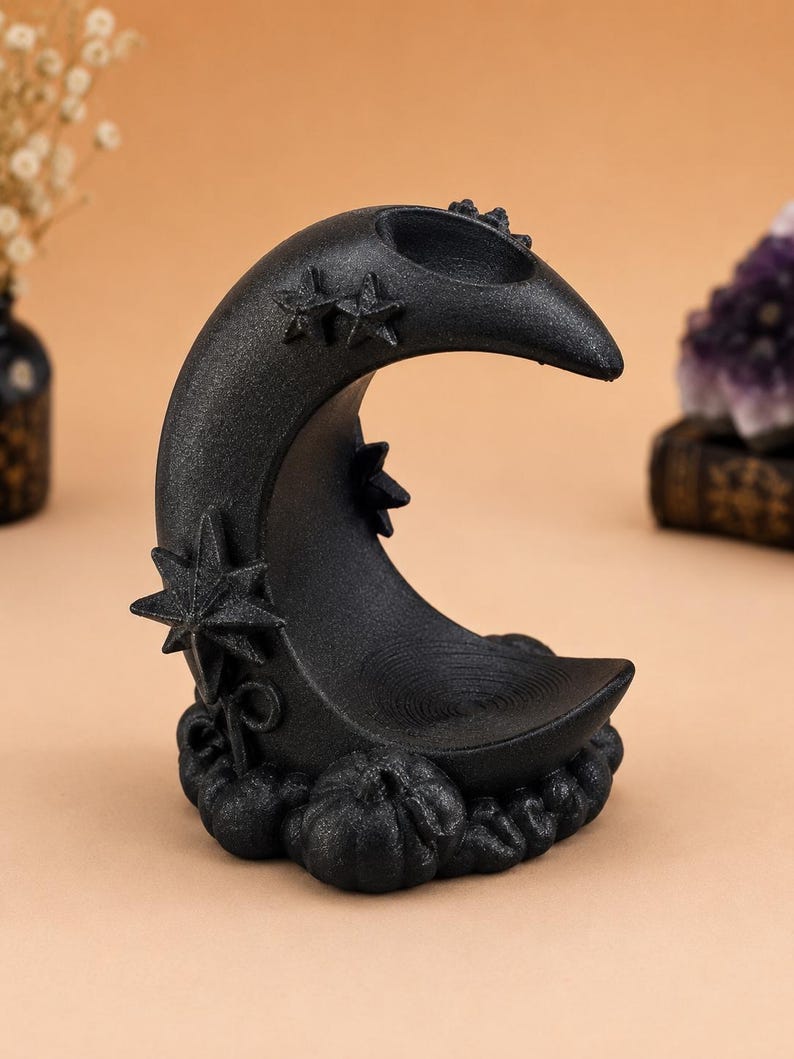 May include: A black crescent moon-shaped incense holder with a textured, glittery finish. The piece features raised star motifs on the curve and a base decorated with small, sculpted pumpkin shapes. A circular indentation sits at the top.