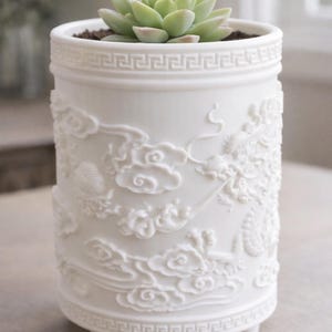 May include: White ceramic planter with a detailed relief design of clouds and a dragon. The cylindrical pot has a Greek key pattern around the top and bottom. A green succulent plant sits inside the planter.