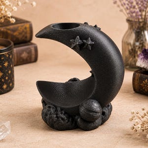 May include: A black crescent moon-shaped incense burner with a textured surface. The design features two small stars on the upper curve and a circular base adorned with spherical planet-like shapes and cloud-like textures at the bottom.