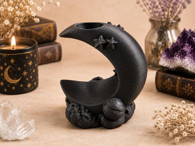 May include: A black crescent moon-shaped incense burner with a textured surface. The design features two small stars on the upper curve and a circular base adorned with spherical planet-like shapes and cloud-like textures at the bottom.