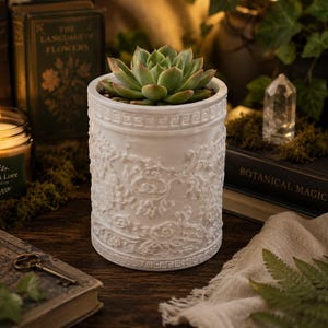 May include: A white cylindrical ceramic planter featuring intricate embossed floral and vine relief patterns on the exterior surface, topped with a green succulent plant. The planter has a decorative geometric border along the top and bottom rims.