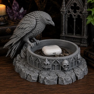 May include: A gray resin trinket tray featuring a sculpted raven perched on the edge. The circular base is decorated with gothic architectural arches and human skull motifs. Inside the tray sits a white wireless earbud charging case.