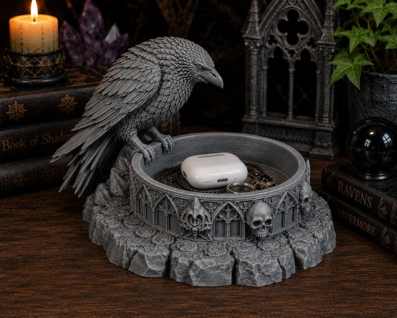 May include: A gray resin trinket tray featuring a sculpted raven perched on the edge. The circular base is decorated with gothic architectural arches and human skull motifs. Inside the tray sits a white wireless earbud charging case.