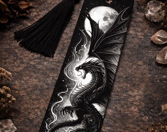May include: Black and white bookmark with a dragon illustration, wings spread against a full moon and starry sky. A black tassel hangs from the top. The design evokes fantasy and gothic themes. The bookmark is a reading accessory.