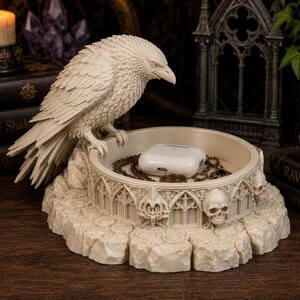 May include: A cream-colored resin trinket dish featuring a sculpted raven perched on the edge. The circular base is decorated with gothic arches and human skulls. Inside the dish sits a white wireless earbud charging case and a gold chain.