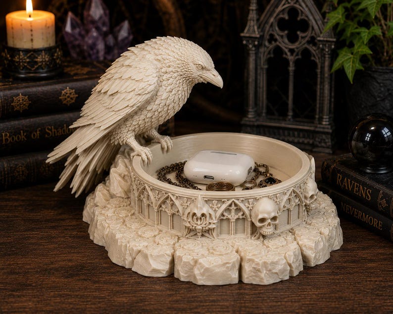 May include: A cream-colored resin trinket dish featuring a sculpted raven perched on the edge. The circular base is decorated with gothic arches and human skulls. Inside the dish sits a white wireless earbud charging case and a gold chain.