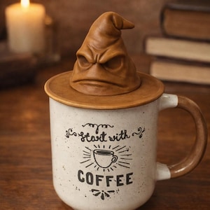 May include: A ceramic mug with a brown lid shaped like a Sorting Hat. The mug is white with brown accents and the words "start with COFFEE" printed on it. A lit candle and books are in the background.