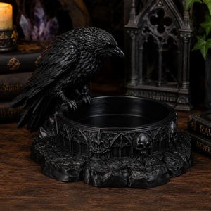 May include: A black raven figurine perched on the edge of a circular bowl. The bowl features a detailed relief pattern of gothic arches and human skulls. The base of the bowl has a textured, rocky appearance, and the entire piece is finished in matte black.