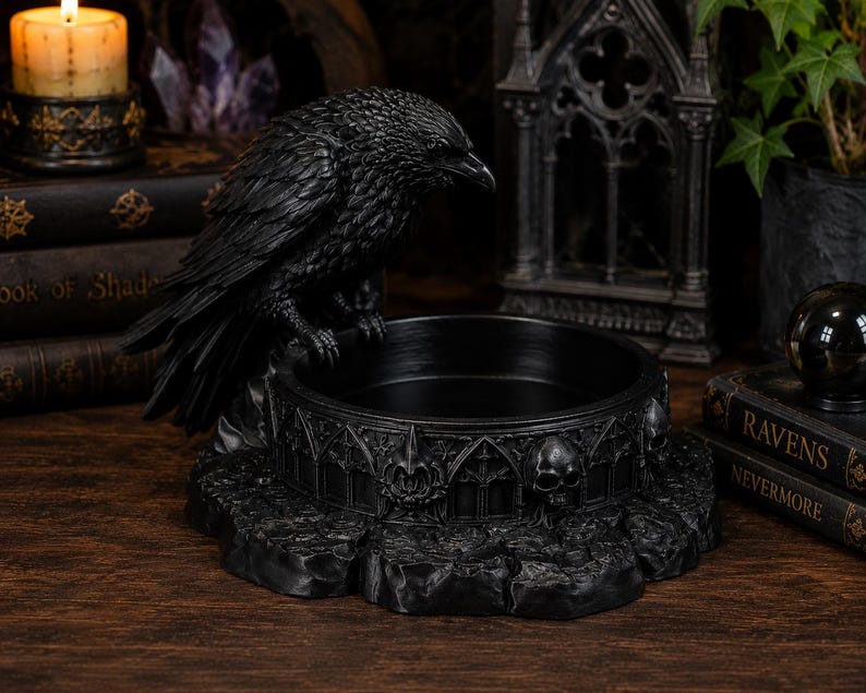 May include: A black raven figurine perched on the edge of a circular bowl. The bowl features a detailed relief pattern of gothic arches and human skulls. The base of the bowl has a textured, rocky appearance, and the entire piece is finished in matte black.