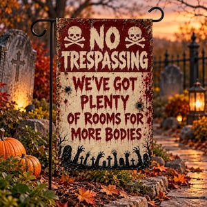 Könnte beinhalten: Eine rechteckige Gartenflagge mit abgenutztem rot-cremefarbenem Hintergrund und zwei Totenköpfen mit gekreuzten Knochen. Der Text lautet NO TRESPASSING WE'VE GOT PLENTY OF ROOMS FOR MORE BODIES in Dunkelrot, ergänzt durch schwarze Silhouetten von Händen und Grabsteinen.