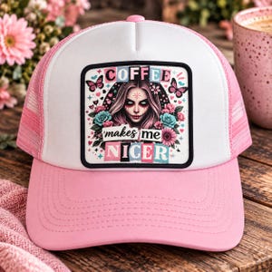 Puede incluir: Una gorra de camionero blanca y rosa con un parche rectangular central que muestra una ilustración gráfica de una persona con cabello largo, rodeada de flores, mariposas y corazones. El texto dice COFFEE makes me NICER en negrita.