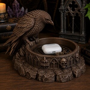 May include: A brown resin trinket tray featuring a sculpted raven perched on the edge. The circular base is decorated with gothic architectural arches and small skull motifs. A white wireless earbud charging case rests inside the tray.