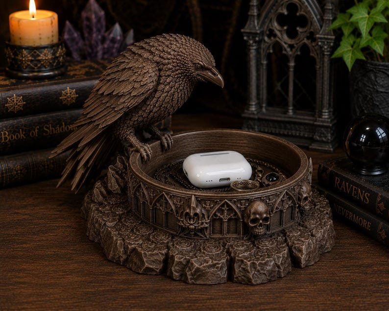 May include: A brown resin trinket tray featuring a sculpted raven perched on the edge. The circular base is decorated with gothic architectural arches and small skull motifs. A white wireless earbud charging case rests inside the tray.