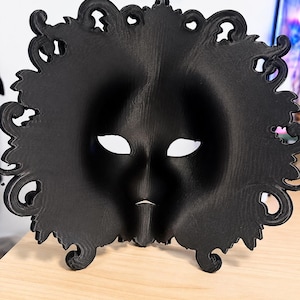 May include: A matte black mask featuring a smooth, contoured face with almond-shaped eye openings and a small, pointed chin detail. The outer edge of the mask is decorated with an intricate, swirling, ornate filigree border design.