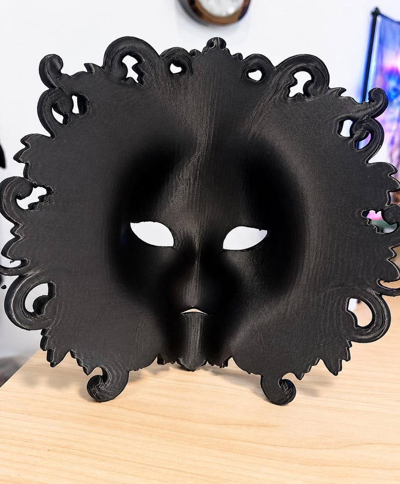 May include: A matte black mask featuring a smooth, contoured face with almond-shaped eye openings and a small, pointed chin detail. The outer edge of the mask is decorated with an intricate, swirling, ornate filigree border design.