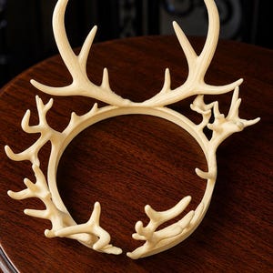May include: Ivory-colored antler crown, featuring a circular base with multiple antler-like projections. The crown is made of a light-colored material and sits on a dark wooden surface. The design is intricate and detailed, resembling a deer's antlers.