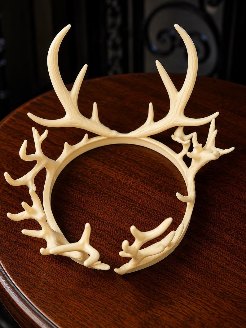 May include: Ivory-colored antler crown, featuring a circular base with multiple antler-like projections. The crown is made of a light-colored material and sits on a dark wooden surface. The design is intricate and detailed, resembling a deer's antlers.