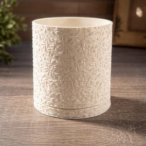May include: A cylindrical, off-white container with a floral embossed pattern. The container is made of a solid material and sits on a matching base. The design features detailed flowers and leaves, creating a textured surface. The container is suitable for holding pencils, makeup brushes, or small items.