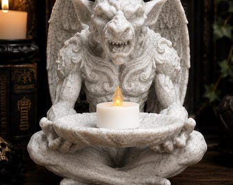 May include: A light gray speckled gargoyle figurine with wings, sitting in a cross-legged position while holding a shallow circular dish. A small white tea light candle with a glowing yellow flame is placed in the center of the dish.