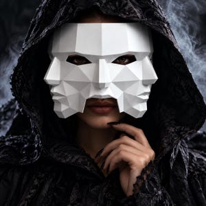May include: A white, geometric, multi-faced mask featuring sharp, angular planes and symmetrical features. The mask covers the upper portion of the face, revealing dark eyes and lips, and is worn with a dark, textured, hooded garment.