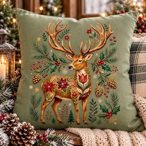 May include: A square sage green throw pillow featuring a central illustration of a deer with large antlers. The deer is decorated with red floral patterns and surrounded by pine branches, red berries, holly leaves, and brown pinecones.