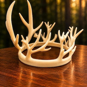 May include: A cream-colored antler crown with multiple points, set on a wooden surface. The crown's design mimics natural antlers, with a circular base and upward-reaching tines. The background is a blurred forest scene.