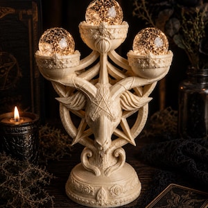May include: A cream-colored resin candle holder featuring a central goat head sculpture integrated into a pentagram design. The structure supports three circular bowls, each containing a glowing, crackled glass sphere light with warm internal illumination.