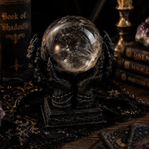 May include: A clear, spherical crystal ball resting atop a black, sculpted base featuring two detailed hands with claw-like fingers. The base is rectangular with intricate embossed patterns, providing a sturdy support for the transparent orb.