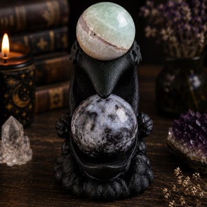 May include: A black raven-shaped sculpture featuring a textured finish, designed to hold two spherical stones. The top stone is a smooth, pale green and white banded sphere, while the bottom stone is a dark grey and black mottled sphere.