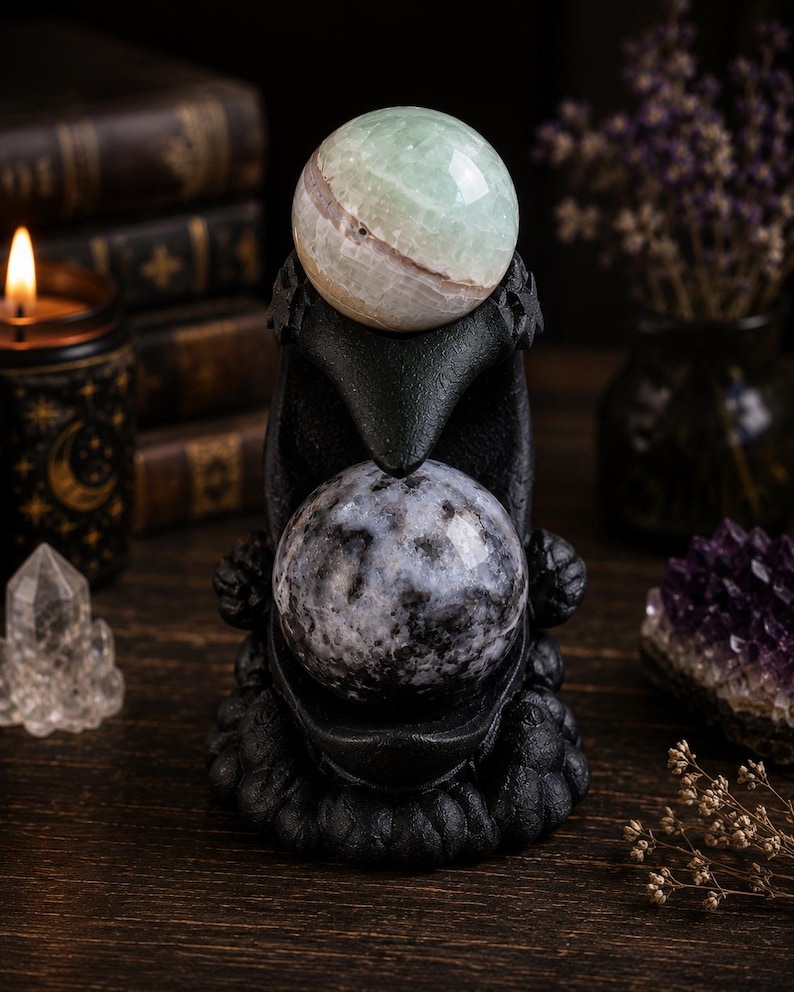 May include: A black raven-shaped sculpture featuring a textured finish, designed to hold two spherical stones. The top stone is a smooth, pale green and white banded sphere, while the bottom stone is a dark grey and black mottled sphere.